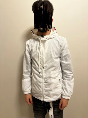 Vit vindjacka från Moncler med huva - Snygg vit vindjacka från Moncler med huva och dragkedja framtill. Jackan har Moncler-logga på ärmen och coola rödblå detaljer på huvan. Långärmad och med snörning vid halsen. Perfekt för dig som gillar stilrena och sportiga plagg.