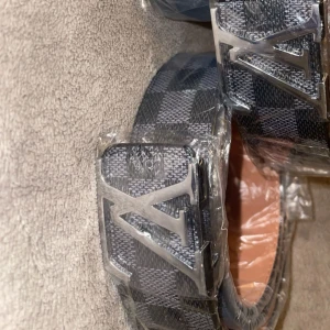  rutig svart bälte - Snyggt bälte från  med klassiskt rutigt mönster i svart och grått. Bältet har en stor metallspänne med logga och är tillverkat i skinn. Perfekt accessoar för att lyfta din outfit.