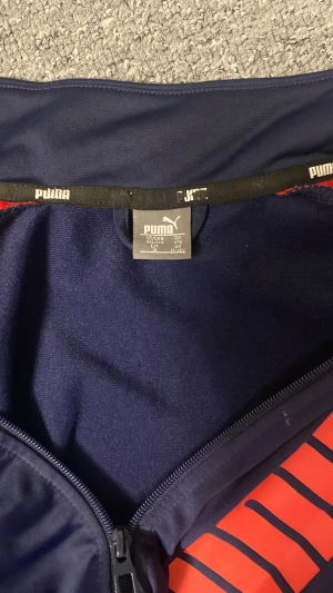 Blå och röd Puma vindjacka - Snygg vindjacka från Puma i mörkblått med röda detaljer längs axlarna och ett stort rött Puma-tryck på framsidan. Jackan har hel dragkedja, hög krage och långa ärmar. Perfekt för dig som gillar sportig stil och vill sticka ut med färg.