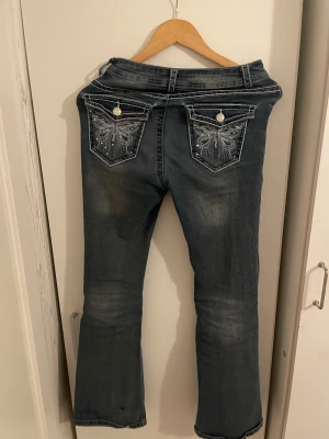 Blå bootcut jeans med fick med knapar och med en fin design av fjärilar  - Snygga blå jeans med bootcut passform och coola broderade bakfickor med fjärilsmotiv och nitar. Tydliga kontrastsömmar och dragkedja framtill. Jeansen har en klassisk femficksdesign och är tillverkade i denim.