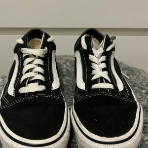Svarta Vans Old Skool sneakers - Klassiska Vans Old Skool sneakers i svart med vit sidorand och vita snören. Skorna har en låg profil, rund tå och platt sula med gummimönster undertill. Ovandelen är i mocka och canvas, med kontrasterande vita sömmar som ger en snygg streetstil.