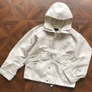 Kaws X The North Face - Säljer en beige regnjacka från The North Face med huva och diskret mönster. Jackan har dragkedja och knappar framtill, två stora fickor och justerbar nederkant. Perfekt för blöta dagar och har en clean streetstil.