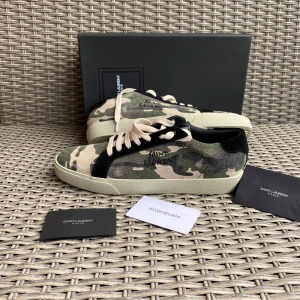 Saint Laurent Court Classic camo sneakers - Snygga Saint Laurent sneakers med camouflage-mönster i grönt, beige och svart. Skorna har vit snörning, platt sula och detaljer i mocka. Saint Laurent-logga broderad på sidan och tryckt på innersulan. Perfekt för dig som gillar streetstyle och exklusiva märken.