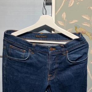  Nudie Jeans Co. Mörkblå - Snygga mörkblå jeans från Nudie Jeans Co med klassisk femficksdesign och orange kontrastsömmar. Modellen har rak passform, knappgylf och läderpatch med logga bak. Perfekta för dig som gillar stilrena och tidlösa jeans. Fråga på bara!