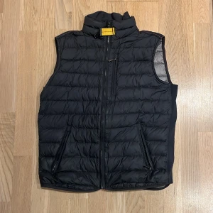 Svart dunväst från Parajumpers - Inga tecken på användning. Svart quiltad dunväst från Parajumpers med ståkrage och gul detalj vid halsen. Västen har två fickor med dragkedja och ett broderat märke på ryggen. Insidan är ljusgrå och materialet är lätt och syntetiskt. Perfekt för lager-på-lager under höst och vinter.