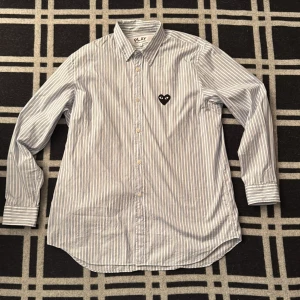 Randig skjorta Comme des Garçons PLAY - Snygg skjorta från Comme des Garçons PLAY i storlek L. Skjortan är vit med blåa ränder och har ett svart hjärta med ögon broderat på bröstet. Klassisk krage, långa ärmar och knappar framtill. Tillverkad i mjuk bomull som känns skön mot huden.