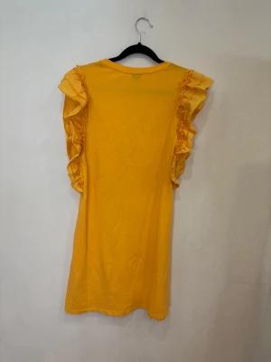 Gul volangklänning från Gina Tricot - Superfin gul kortklänning från Gina Tricot med volangdetaljer längs axlarna. Klänningen har rund halsringning och är ärmlös med snygga ryschade volanger som ger en cool och lekfull look. Perfekt för sommarens alla tillfällen!