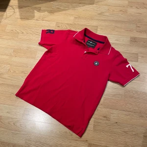 Röd Sail Racing pikétröja  - Riktigt snygg röd pikétröja från Sail Racing med vit piping på krage och ärmslut. Broderad logga på bröstet och tryckta siffror på ärmarna. Klassisk passform och mjukt bomullsmaterial som känns skönt mot huden. Perfekt för dig som gillar sportigstil. Det är välldigt bra skick. Tröjan är i storlek 160