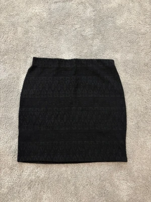 Svart kortkjol med diskret mönster - Svart kortkjol med diskret geometriskt mönster i tyget. Kjolen har en rak och tight passform som sitter snyggt på kroppen. Materialet känns mjukt och har lite struktur, perfekt för dig som gillar stilrena plagg med subtila detaljer.