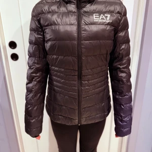 Svart dunjacka EA7 Emporio Armani - Svart dunjacka från EA7 Emporio Armani med stilren design och logga på bröstet. Jackan har quiltade paneler, hel dragkedja framtill och långärmad passform. Perfekt för kyliga dagar och har en lätt, smidig känsla. Snyggt märke på dragkedjan och bröstet.