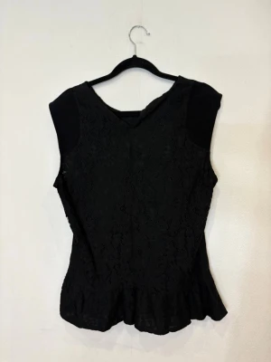 Svart spetsblus med volangkant - Säljer en svart blus med spetsdetaljer och volangkant nertill. Blusen är ärmlös och har en rund halsringning. Perfekt för dig som gillar en stilren men ändå romantisk look. Materialet är mjukt och följsamt med vacker spets över hela framsidan.