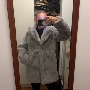 Grå pälsjacka från Vamiia - Säljer en fluffig och grå pälsjacka från Vamiia i storlek M. Jackan har långa ärmar och är helfodrad. Perfekt för dig som vill ha en mysig och trendig look under kalla dagar. Jackan har en stilren design med knappar men som är osynliga.