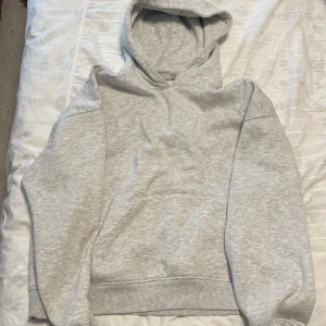 Ljusgrå hoodie med fleecefoder från Gina Tricot - Mjuk och skön ljusgrå hoodie med huva och ribbade muddar. Insidan är fodrad med fluffig fleece som gör den extra varm och bekväm. Tröjan har en klassisk passform och är perfekt för chill dagar. Enkel design utan tryck eller mönster.