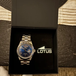 Lotus herrklocka med blå urtavla - Snygg herrklocka från Lotus med blå urtavla och dag- och datumvisning. Klockan har en robust länk i silverfärgat stål och markerad boett med räfflad kant. Klassisk och stilren design som passar perfekt till jeans eller skjorta.