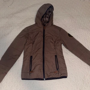 Brun pufferjacka från Jacson med huva - Snygg brun pufferjacka från Jacson med huva och svarta detaljer. Jackan har två fickor med dragkedja, emblem på ärmen och slät, glansig yta. Perfekt för dig som gillar stilren och sportig look. Finns i storlek S.