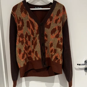 Brun leopardmönstrad kofta Acne Studios - Snygg brun kofta från Acne Studios med leopardmönster i bruna och beige toner på framsidan. V-ringad med knappar hela vägen och ribbade muddar. Perfekt för dig som vill sticka ut med ett coolt mönster och skön passform.