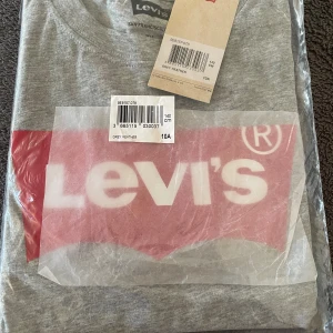 Grå Levi's t-shirt med logga - Snygg grå t-shirt från Levi's med klassisk röd och vit logga framtill. T-shirten har rund hals och är kortärmad, perfekt för en avslappnad och cool stil. Tillverkad i mjuk bomull för maximal komfort.