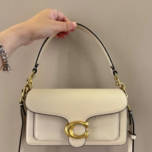 Coach axelväska i skinn - Stilren axelväska från Coach i ljusbeige skinn med grov struktur. Väskan har ett stort guldfärgat C-spänne framtill, avtagbar axelrem och detaljer i guldmetall. Perfekt storlek för det viktigaste och en klassisk look.