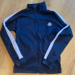 Blå vindjacka från Moncler, strl S - Snygg marinblå vindjacka från Moncler med vita ränder längs ärmarna och klassisk logga på bröstet. Jackan har hel dragkedja framtill, hög krage och ribbade muddar. Perfekt för dig som gillar sportig och clean stil.