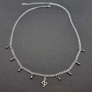 Silverfärgat halsband med musikberlock - Stilrent halsband i silverfärg med små hängande berlocker och en större berlock i form av en g-klav. Kedjan är tunn och har justerbar längd. Perfekt accessoar för dig som älskar musik och vill ha något unikt och coolt. Material-rostfritt stål 