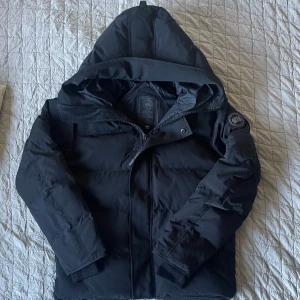 Canada Goose MacMillan Black Badge - Säljer en Black Badge Canada Goose jacka. Jackan har huva, broderad logotyp på ärmen och dragkedja framtill. Materialet är högt kvalité och dun, perfekt för kalla dagar. Stilren design med mörka detaljer och klassisk Canada Goose-märke.