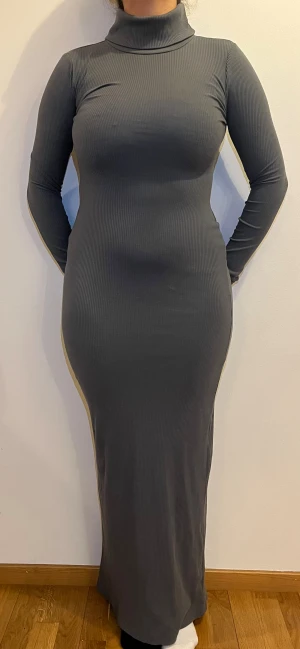 ribbad klänning - Säljer en snygg grå långklänning med polokrage och långärmade ärmar. Klänningen är ribbad och har en tight passform som framhäver figuren. Baktill finns en slits för extra rörelsefrihet. Perfekt för dig som gillar minimalistisk och stilren look. Storlek: EU: 38 UK:10 US:6  Använd 1 gång! Som ny!  Material:  62% polyester, 34% bomull 4%elastan   Obs! Modellens längd är 162 (så blir som en maxi klänning)