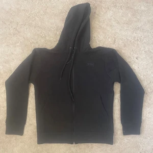Svart quiltad hoodie från BOSS - Svart hoodie från BOSS med quiltat rutmönster, dragkedja framtill och snörning i huvan. Tröjan har långa ärmar, två fickor och en stilren BOSS-logga på bröstet. Perfekt för dig som gillar en clean och modern look.