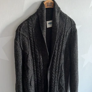 Mörkgrå stickad kofta Old Navy - Mysig mörkgrå stickad kofta från Old Navy med grova kabelstickade detaljer och bred krage. Koftan har knappar framtill och ett chunky, strukturerat mönster som ger en cool och avslappnad vibe. Perfekt att slänga över en t-shirt när det är kyligt ute.