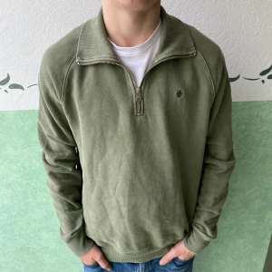 Olivgrön half zip tröja Morris - Snygg olivgrön tröja från Morris med half zip och hög krage. Tröjan har ett litet broderat emblem på bröstet och är tillverkad i mjuk bomull. Perfekt för dig som gillar stilrena och bekväma plagg med lite sportig vibe.