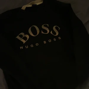 Svart sweatshirt från Hugo Boss - Svart sweatshirt från Hugo Boss med stort, guldigt BOSS-tryck på bröstet. Tröjan har rund hals och långa ärmar, perfekt för dig som gillar stilrena och klassiska märkesplagg. Materialet känns mjukt och bekvämt mot huden.