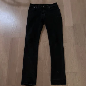 Svarta jeans från Nudie Jeans - Snygga svarta jeans från Nudie Jeans med klassisk femficksdesign och orangea detaljer på bakfickan och midjelappen. Jeansen har rak passform och är tillverkade i slitstarkt denimtyg. Perfekta för dig som gillar stilrena och tidlösa jeans.