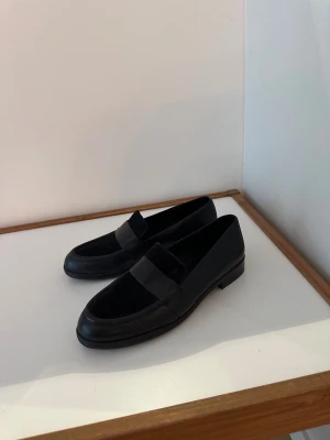 Svarta loafers i skinn och mocka - Snygga svarta loafers från Other Stories med klassisk design. Ovandelen är i mjukt skinn med detaljer i mocka och har en rund tå samt platt sula. Perfekta för dig som gillar stilrena och tidlösa skor med en modern touch. Jag har storlek 39/40 och de är något för små för mig.