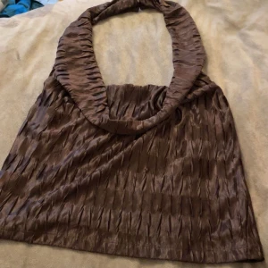 Brun omlott halterneck topp - Säljer en brun halterneck topp med omlottkrage och snyggt veckat tyg. Toppen har en glansig finish och är perfekt för dig som gillar en lite lyxigare känsla. Passformen är loose och materialet känns som satin eller polyester. Pris kan diskuteras och köparen står för frakt