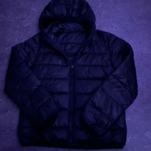 Svart pufferjacka från Champion  - Svart pufferjacka från Champion i ålder 13-14 (162cm-167). Jackan har huva, dragkedja framtill och quiltad design. Materialet är glansigt och syntetiskt, perfekt för kalla dagar. Klassisk look med långärmade ärmar och diskret logga på insidan av huvan.
