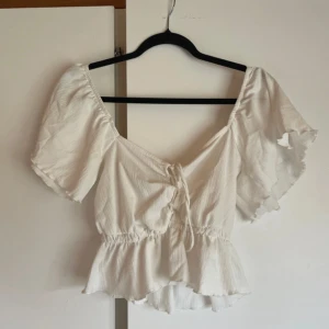 Vit omlott blus med volangärm - Supersöt vit blus med omlottkänsla och snörning framtill. Toppen har korta, volangiga ärmar och resår i midjan för en snygg passform. Perfekt till jeans eller kjol för en fräsch look. Materialet är lätt och luftigt, perfekt för varmare dagar.