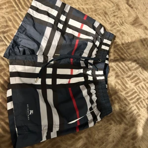 Randiga badshorts från Burberry - Snygga badshorts från Burberry med klassiskt randigt mönster i svart, vit, grå och röd. Shortsen har elastisk midja med snörning och meshfoder på insidan. Perfekta för stranden eller poolen, med stilren Burberry-logga på benet.