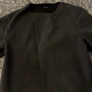 Mörkgrå kappa  - Snygg mörkgrå kappa från Gina tricot. Jackan har en enkel knapp framtill och är tillverkad i mjukt ullmaterial.