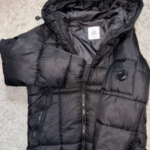 Svart pufferjacka från C.P. Company - Säljer en svart pufferjacka från C.P. Company med ikonisk linsdetalj på ärmen. Jackan har huva, två sidofickor med dragkedja och quiltad design. Perfekt för kalla dagar och har en modern, clean look som funkar till streetwear.