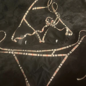 Svart bikini med rutiga band, strl L - Snygg svart bikini med klassiska rutiga band i beige, svart och rött. Toppen har trekantsform och knyt i nacken och ryggen, medan trosan har smala band på sidorna. Materialet är 80% polyester och 20% elastan, perfekt för sommaren och strandhäng.