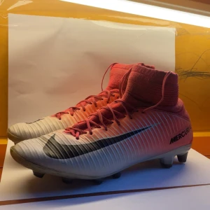 Nike Mercurial fotbollsskor rosa/vit - Nike Mercurial fotbollsskor med högt skaft i en snygg gradient från rosa till vit. Skorna har svarta Nike-loggor, snörning och platt sula med dobbar för gräsplan. Ovandelen är tillverkad i syntetmaterial och har en tight passform för maximal kontroll på planen.