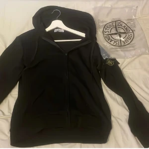 Svart hoodie från Stone Island M - Svart hoodie från Stone Island med klassisk logotyp på ärmen. Tröjan har huva, hel dragkedja framtill och långa ärmar. Priset kan diskuteras vid riktigt snabb affär! Riktigt fet nu på vintern 