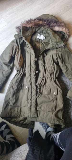 Olivgrön parkajacka med fuskpäls - Parkajacka i olivgrönt med stor huva kantad av fuskpäls. Jackan har dragkedja och knappar framtill, snörning i midjan och två stora fickor med lock. Insidan är fodrad med mjuk teddy för extra värme. Perfekt för kalla dagar.