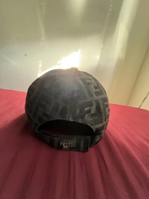 Svart Fendi keps med FF-mönster - Snygg svart keps från Fendi med det klassiska FF-logomönstret över hela kepsen. Kepsen har böjd skärm med svart kant och justerbar rem baktill med Fendi-logga. Materialet är mjukt och känns lyxigt, perfekt för dig som gillar designeraccessoarer.