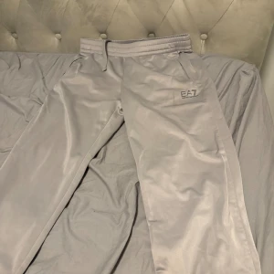 Grå EA7 byxor med snörning - Snygga grå byxor från EA7 med elastisk midja och snörning för perfekt passform. Materialet är blankt och känns mjukt mot huden, med coola svarta detaljer på benet. Perfekta för träning eller chill. EA7-logga framtill ger en sportig vibe.