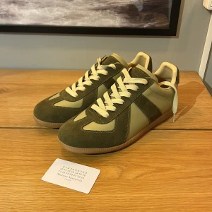 Maison Margiela sneakers i olivgrönt - Maison Margiela sneakers i olivgrönt och beige med snygga kontraster mellan mocka och skinn. Klassisk rund tå, platt gummisula och vita snören. Ikonisk design med märkning på sula och insida. Perfekt för dig som gillar stilrena och unika sneakers.