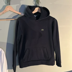 Mörkblå hoodie från Lacoste med logga - Mörkblå  hoodie från Lacoste med klassisk krokodillogga på bröstet. Tröjan har huva, känguruficka och ribbade muddar. Tröjan köpte jag för några månader sen. Ganska bra skick bara använd några gånger. Säljer då den blivit för liten för mig. Hör av dig vid fler frågor:)