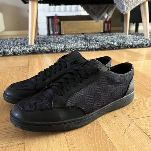 Louis Vuitton svarta sneakers damier - Svarta Louis Vuitton sneakers med klassiskt damier-rutmönster på sidan och ovandelen. Skorna har svarta snören, rund tå och platt sula med LV-logga undertill. Materialmix av skinn och textil ger en lyxig känsla och stilren look. Riktigt bra skick 