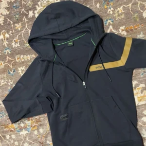 Svart hoodie från BOSS med guldtryck - Snygg svart hoodie från BOSS med dragkedja och huva. Guldigt tryck och logga på ena ärmen ger en exklusiv vibe. Grön detalj på insidan av kragen. Perfekt för dig som gillar streetwear och vill sticka ut med stil.