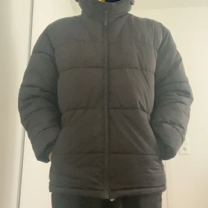 Svart pufferjacka med huva - Svart pufferjacka med huva och dragkedja framtill. Jackan har en quiltad design med breda paneler och långärmade ärmar. Passformen är normal och materialet känns som syntet, perfekt för kalla dagar. Enkel och stilren look som funkar till det mesta.