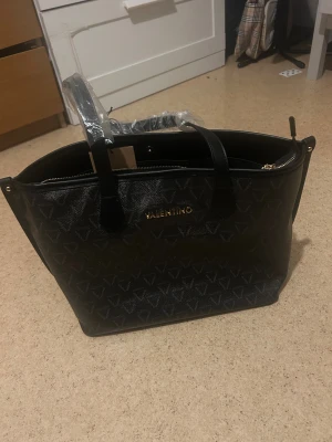 Svart handväska från Valentino - Stilren svart handväska från Valentino med subtilt mönster av V-loggor över hela väskan. Väskan har guldfärgade detaljer, två handtag och dragkedja upptill. Perfekt storlek för att få plats med allt du behöver och passar till många olika looks.